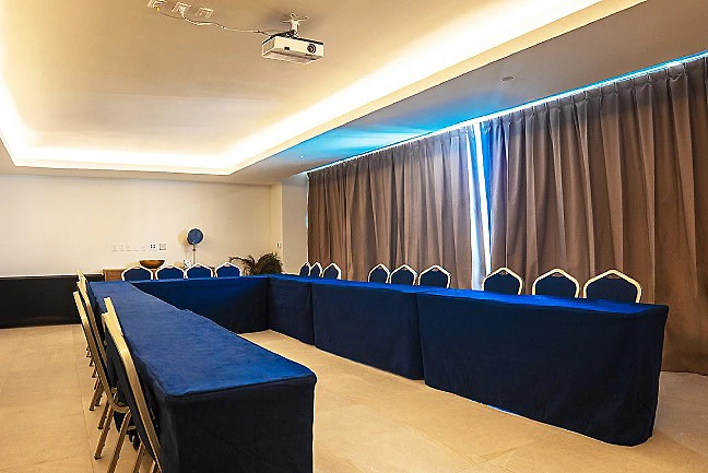 Sala de conferencias con mesas azules y sillas, bajo luz tenue.