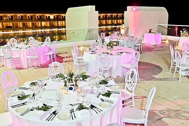 Mesa de banquete con sillas blancas y decoraci&oacute;n rosa, en terraza iluminada