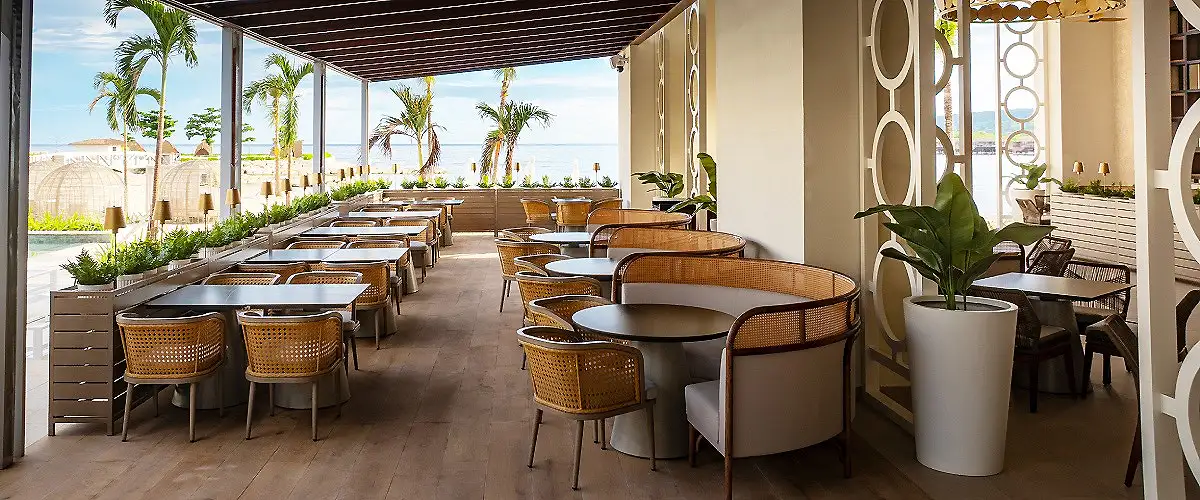 Terraza con mesas, sillas y palmeras frente al mar, con ambiente tropical.