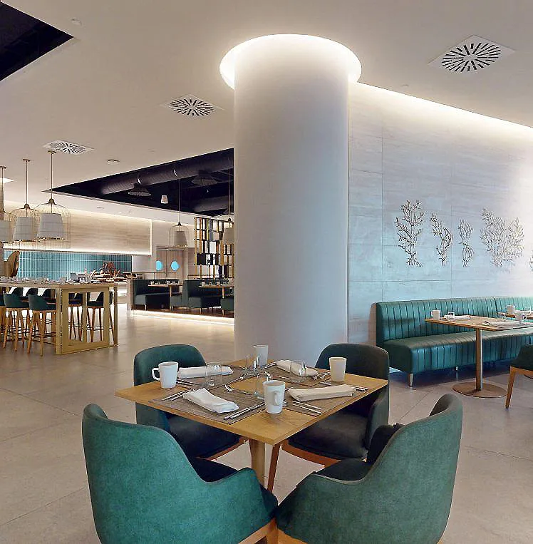 Interior de hotel con mesas, sillas verdes y barra al fondo, luz c&aacute;lida