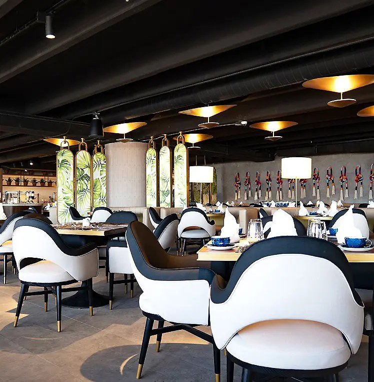 Comedor elegante con mesas, sillas blancas y l&aacute;mparas colgantes, barra al fondo.