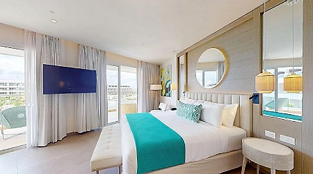 Habitaci&oacute;n de hotel con cama blanca y colcha turquesa, ventana y TV
