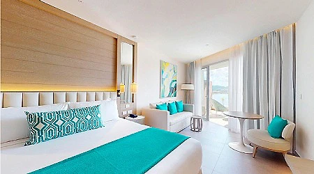 Habitaci&oacute;n de hotel luminosa con cama, sill&oacute;n azul y mesa junto a ventana