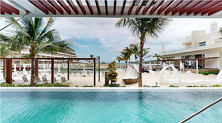 Piscina al aire libre con palmeras y zona de descanso junto al mar.