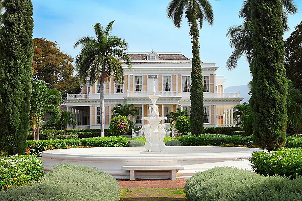 Casa colonial blanca con fuente y jardines, palmeras y cipreses.