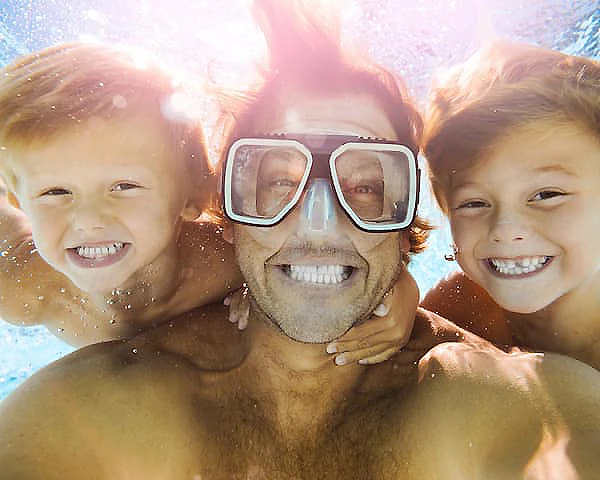 Hombre y dos ni&ntilde;os sonr&iacute;en en el agua con gafas de sol flotando