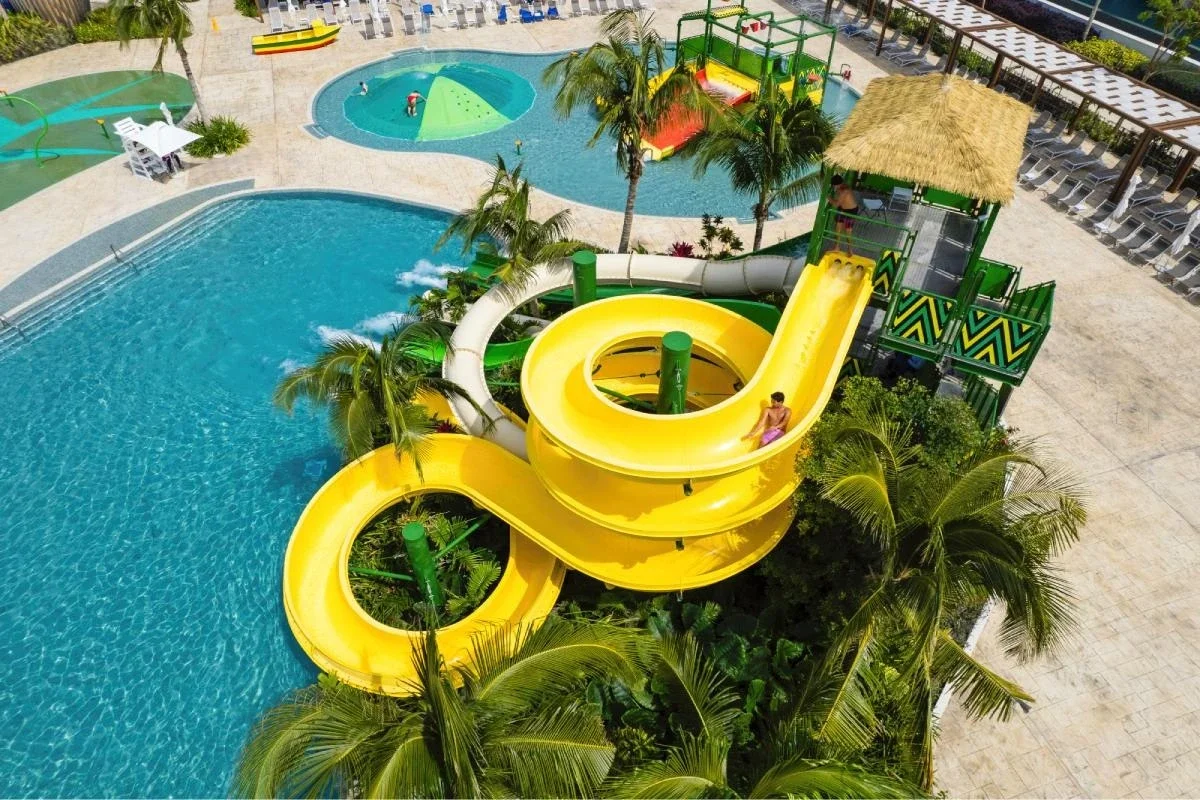 Parque acu&aacute;tico con toboganes amarillos y piscinas, palmeras y zona infantil