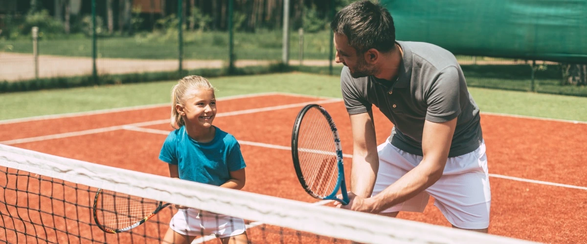 Padre y ni&ntilde;a juegan tenis en una pista de arcilla, raquetas en mano.