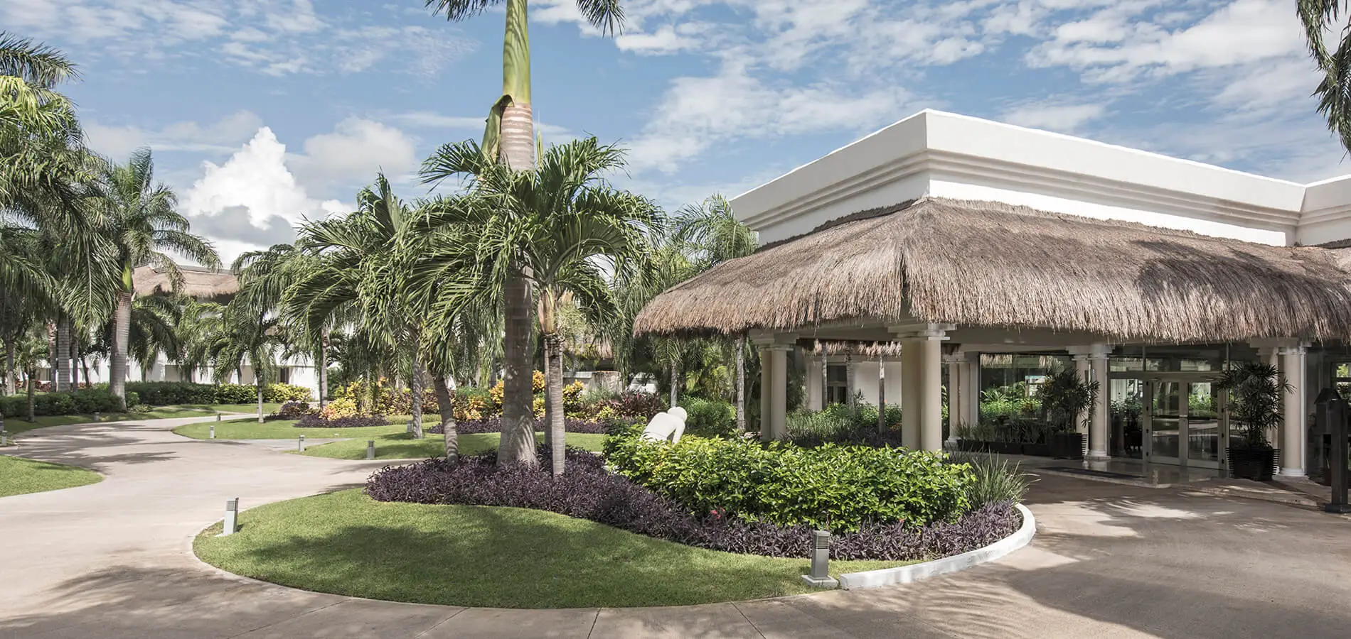 Jard&iacute;n tropical con palmeras y caba&ntilde;a de techo de paja junto a camino.