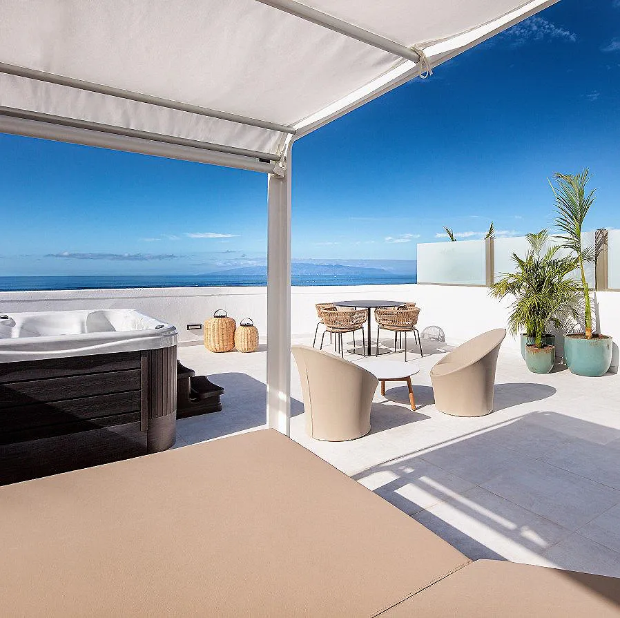 Terraza frente al mar con jacuzzi, mesa y sillas bajo toldo, palmeras.