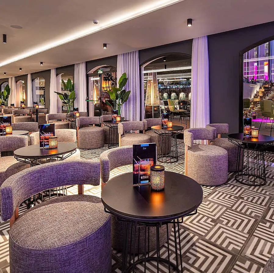 Sal&oacute;n de hotel con mesas, sillas grises, velas luminosas y cortinas blancas.