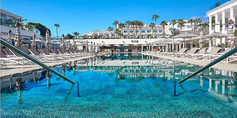 Piscina azul con tumbonas y palmeras bajo un cielo despejado, junto a un hotel.