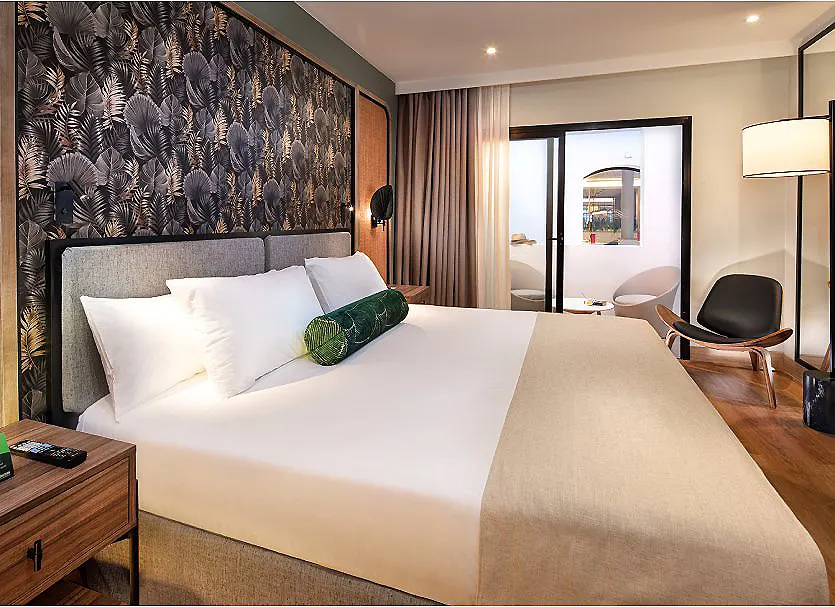 Habitaci&oacute;n de hotel moderna con cama, cortinas y mesa lateral.