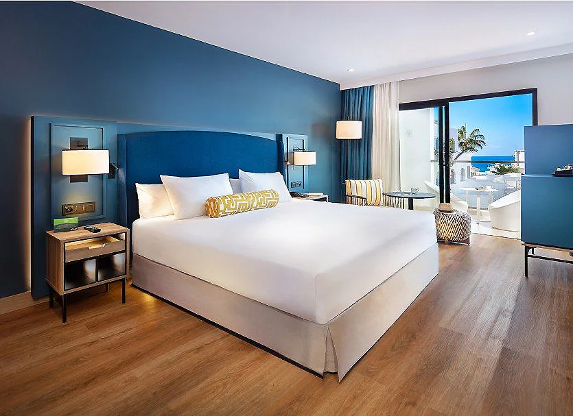Habitaci&oacute;n hotelera moderna con cama blanca y paredes azules, vista al mar