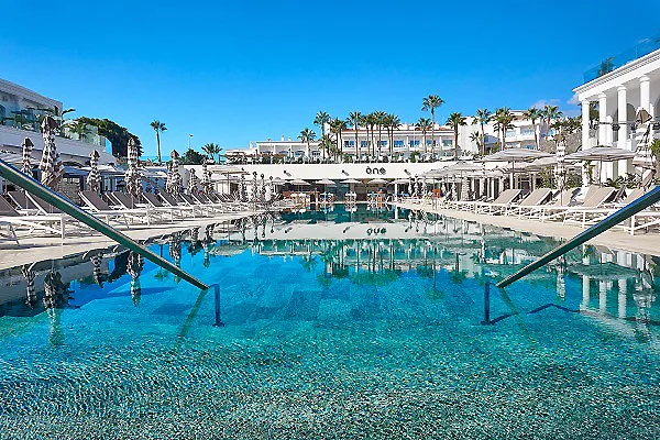 Piscina con palmeras y tumbonas junto al mar, en un complejo tur&iacute;stico.
