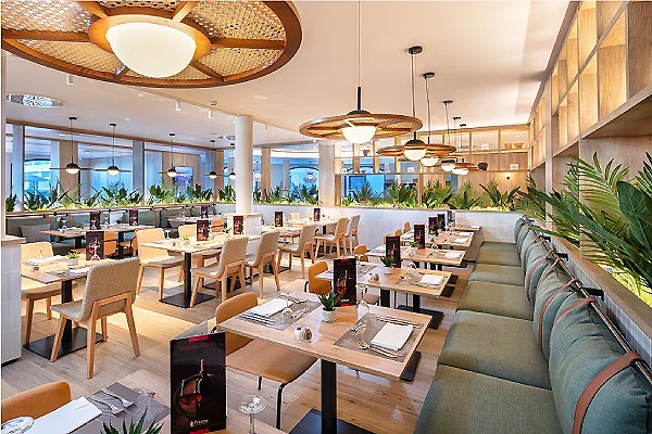 Interior de restaurante con mesas, iluminaci&oacute;n y plantas junto a ventanales.