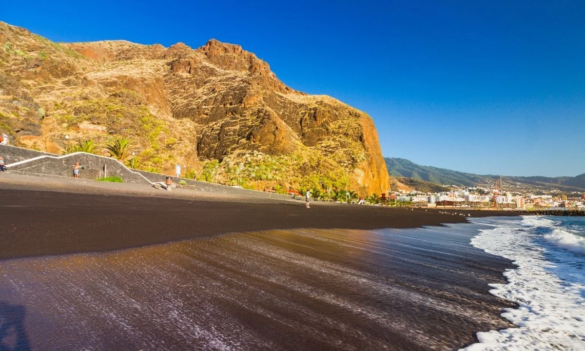 Shoreline at Playa de Bajamar showing the contrast of white sea foam against the extensive black volcanic sand, flanked by a huge rocky cliff under a clear sky.