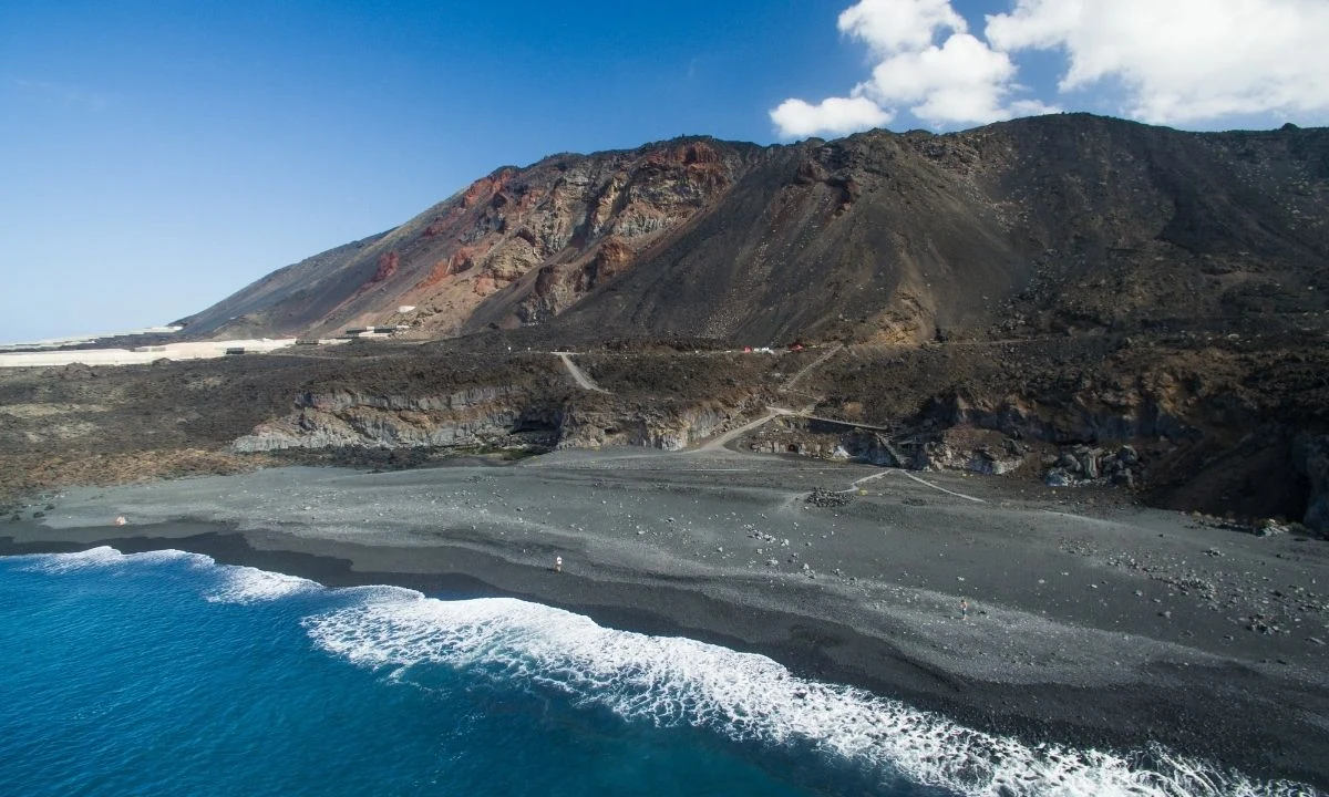 Aerial view of Playa de Echentive with its black sand and pic&oacute;n shoreline, surrounded by an expansive, arid volcanic landscape of lava flows under a blue sky.
