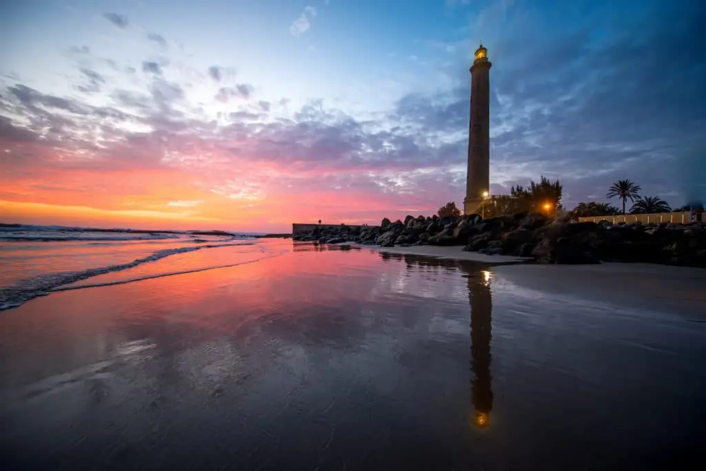 Atardecer con faro iluminado reflejado en la playa y el mar.