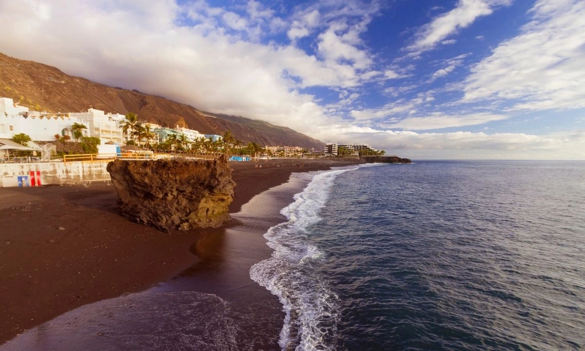Playa de Puerto Naos with its characteristic black volcanic sand, rocks along the shore, coastal buildings and palm trees under a partly cloudy sky in La Palma. 