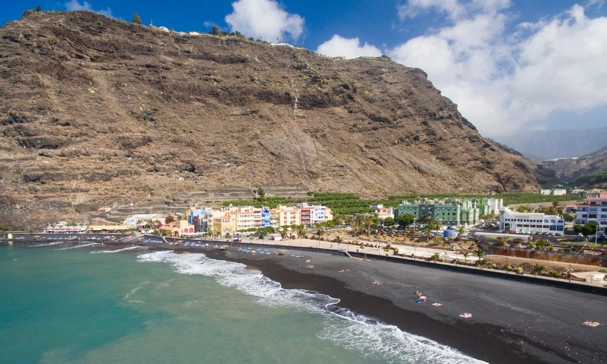 Aerial panorama of Playa de Tazacorte with parasols on its black sand, calm turquoise waters and the colourful buildings of the town set against a large cliff.