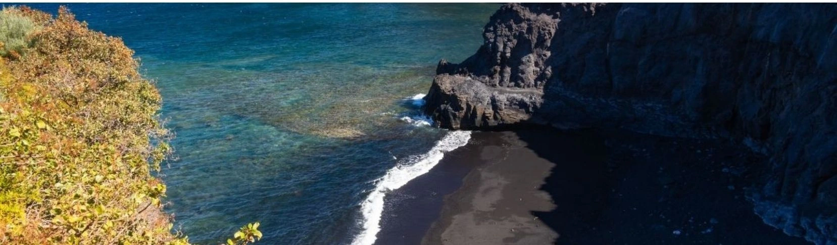 View from above of Playa de La Zamora in Fuencaliente, showing a black sand cove surrounded by imposing dark cliffs and rock formations in the blue sea.