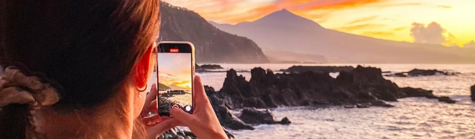 Persona con m&oacute;vil fotografiando un atardecer en la costa y monta&ntilde;as al fondo