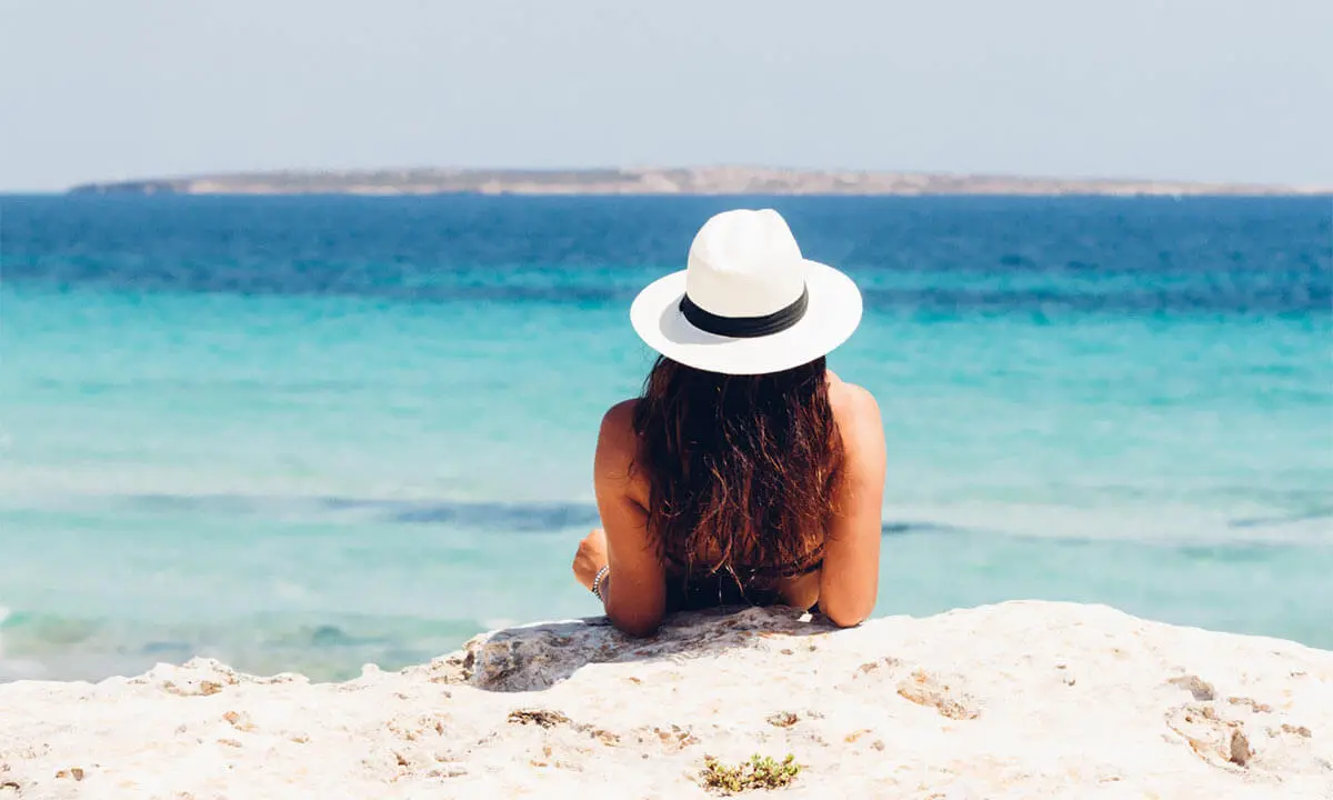 Mujer con sombrero blanco frente al mar turquesa desde una roca