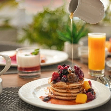 Pancakes con frutas rojas y miel, servidos al aire libre junto a jugo.
