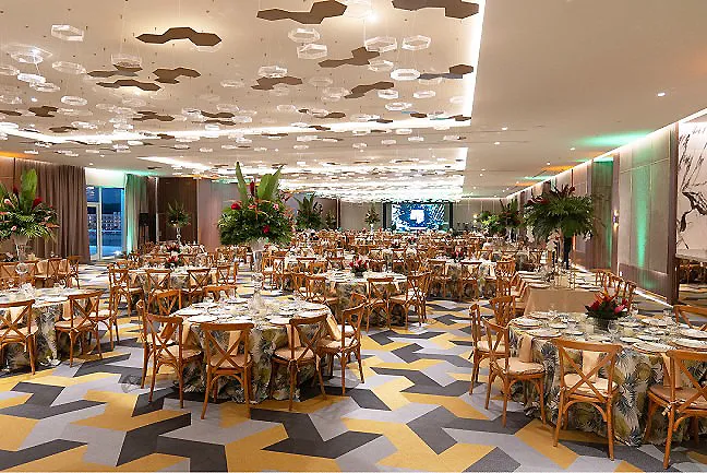 Sal&oacute;n de eventos con mesas y sillas, plantas decorativas y luz natural.