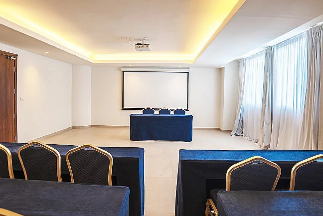 Sala de conferencias con pantalla y butacas azules, luces c&aacute;lidas y cortinas.