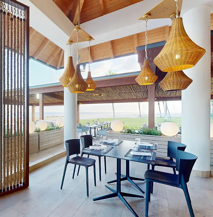 Comedor con mesa azul, l&aacute;mparas de mimbre y techo de madera junto al mar.