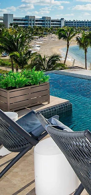 Vista de piscina con palmeras y hotel junto a la playa.