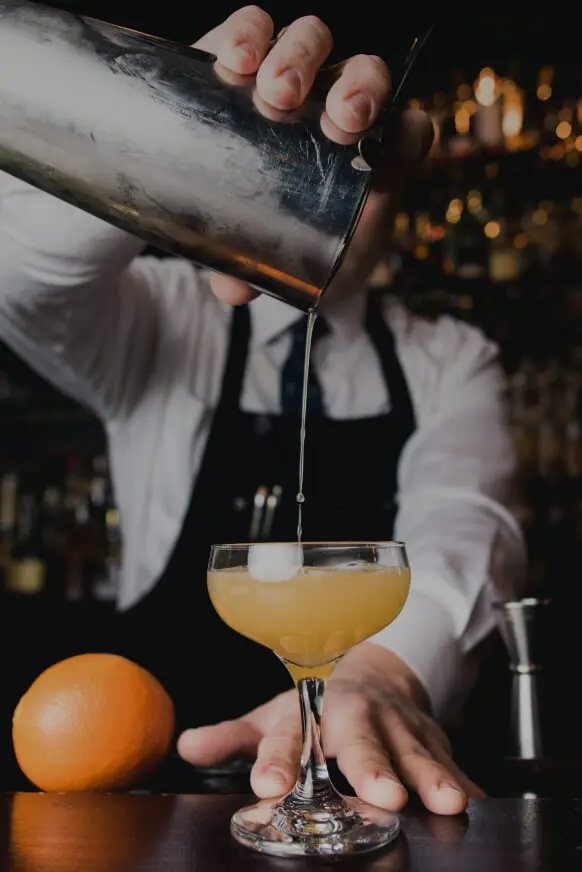 Bartender sirve un c&oacute;ctel con hielo, naranjas y luces de fondo nocturnas.