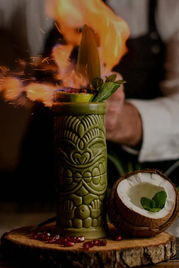 Copa verde tallada con hojas y coco, con fuego y salpicaduras al fondo.