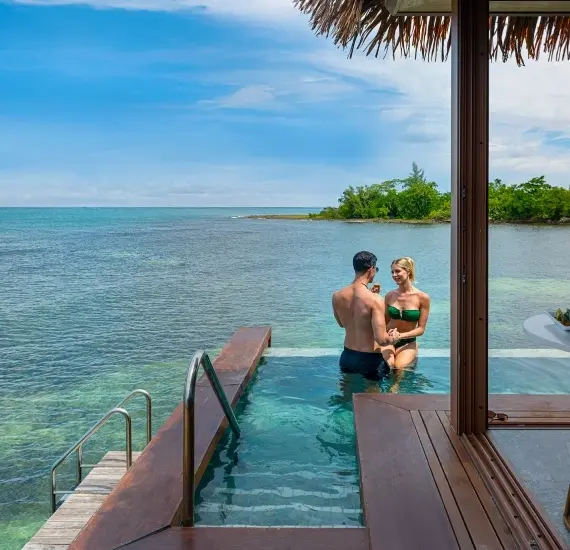 Pareja en una piscina frente al mar, con playa y vegetaci&oacute;n al fondo.