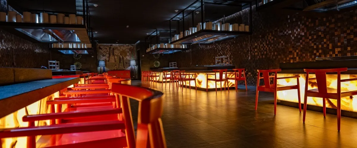 Interior de bar con mesas y sillas rojas, luces c&aacute;lidas y estanter&iacute;as.