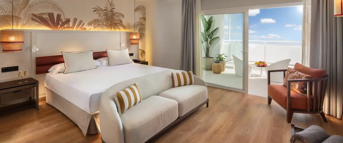 Dormitorio moderno con cama doble, sill&oacute;n y luz natural junto al balc&oacute;n.
