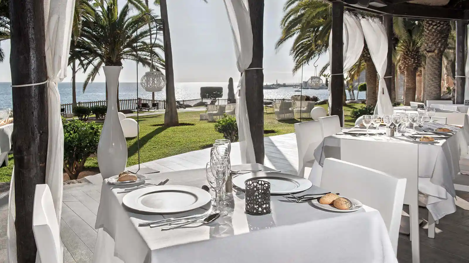 Mesa de banquete con manteles blancos junto al mar, palmeras y jard&iacute;n