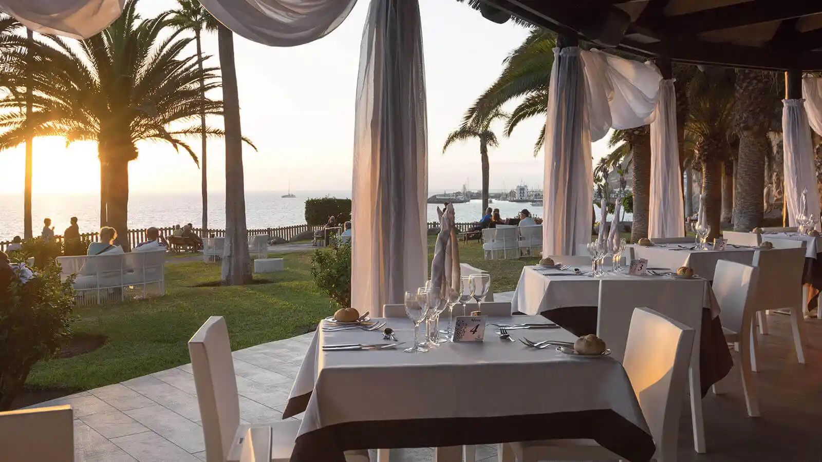 Cena al aire libre en jard&iacute;n junto al mar, mesas con manteles blancos y palmeras al atardecer