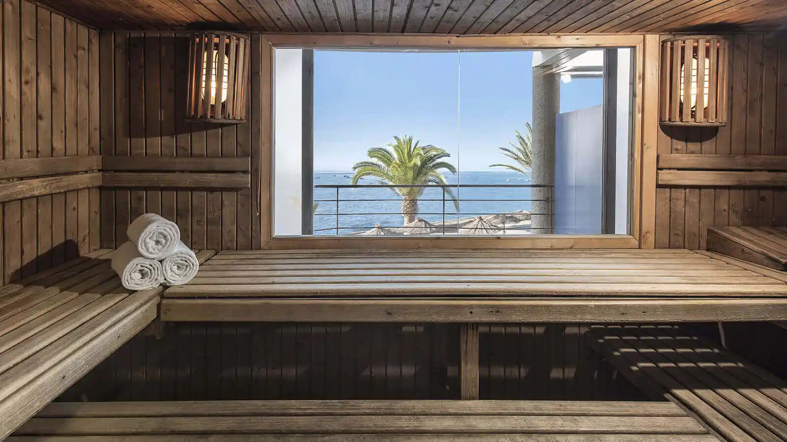 Sauna de madera con toallas enrolladas y ventana al mar y palmeras