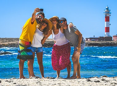 Familia sonriendo en la playa, frente a un faro y el mar azul.