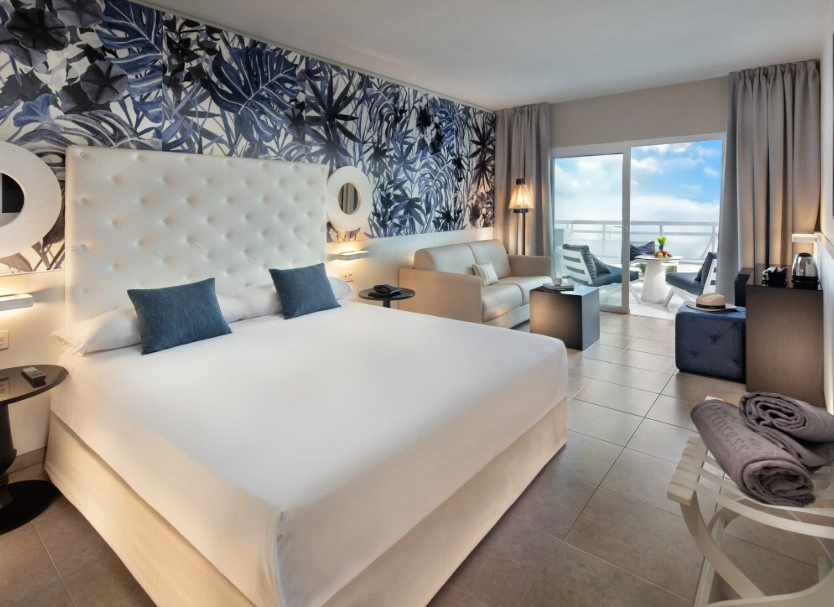 Dormitorio moderno con cama, cortinas grises y vista al mar desde la ventana.