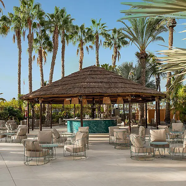 Gazebo de palapa con mesas y sillas junto a palmeras y piscina.