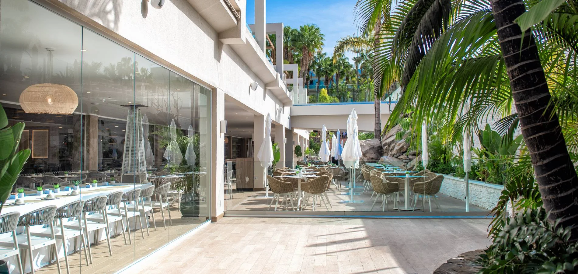 Terraza hotelera con mesa, sombrillas y palmeras junto a un comedor al aire libre