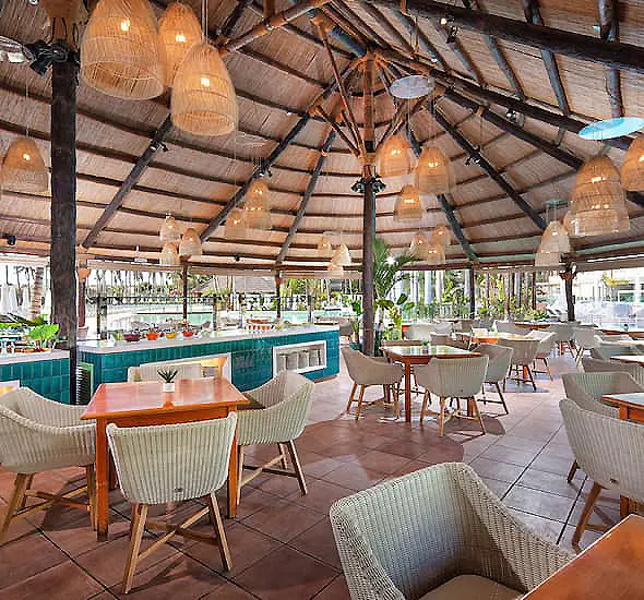 Interior de un restaurante tropical con mesas, sillas y l&aacute;mparas colgantes.