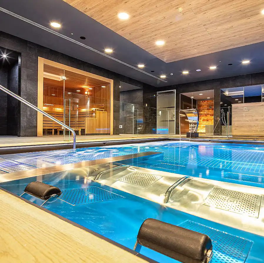 Piscina cubierta moderna con luz LED, piso de madera y zona de bienestar con asientos