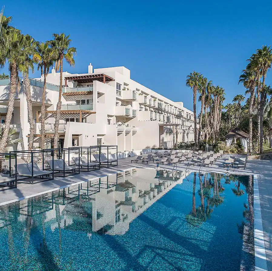 Hotel blanco con piscina y palmeras bajo cielo azul, vista lateral.