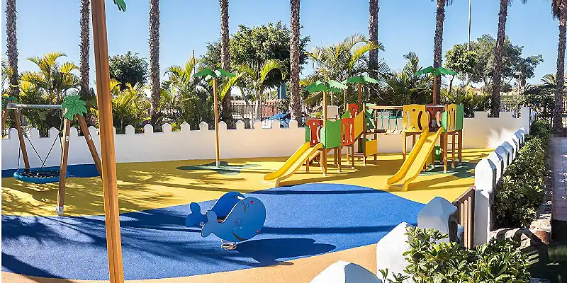 Parque infantil con toboganes, piscina infantil y palmeras al fondo.