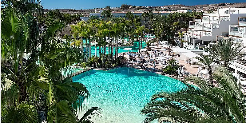Piscina turquesa con palmeras y complejo hotelero al fondo.